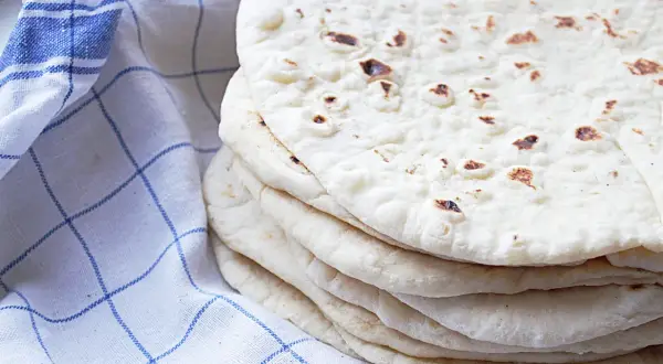 En hög med nybakade tortillabröd, perfekta för ditt nästa tortilla recept. Bredvid ligger en blåvitrutig kökshandduk.