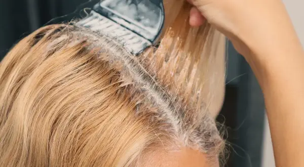 Ręka z pędzlem nakłada produkt do rozjaśniania włosów w domu. Widać blond pasma i początek zabiegu.