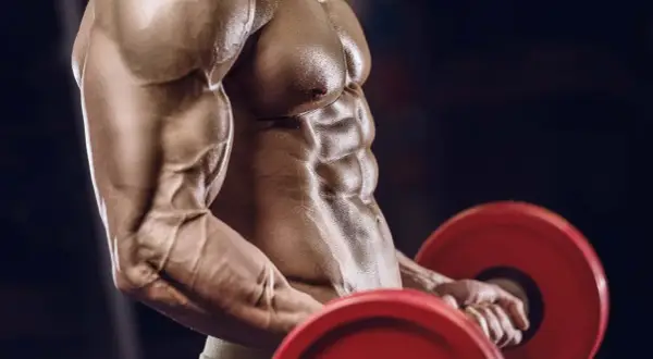 Ein Mann mit stark definierten Bauchmuskeln und Armen hebt eine Hantel. Die Muskelpumpe ist deutlich sichtbar, ein Zeichen intensiven Trainings.