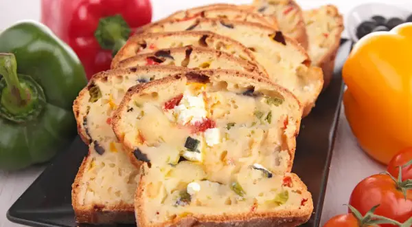 Tranches d'un cake savoureux aux olives et feta, entouré de poivrons colorés et de tomates.