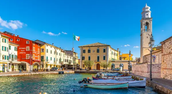 Colorful buildings line a harbor with boats, an Italian flag, and a bell tower under a clear blue sky.