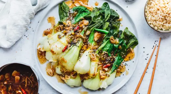 Fertig zubereiteter Pak Choi mit einer würzigen Sauce, garniert mit Sesam und Chili. Dazu gibt es Reis und Stäbchen.