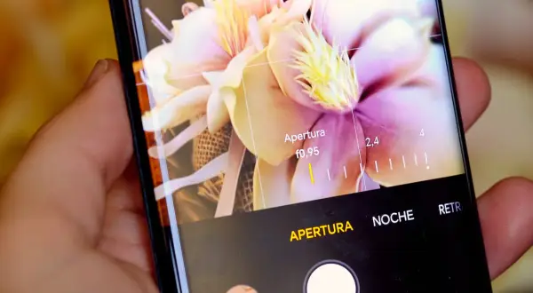 Mano sujetando móvil, mostrando interfaz de cámara con flores y ajustes de apertura. Aprende como hacer un selfie con el móvil.