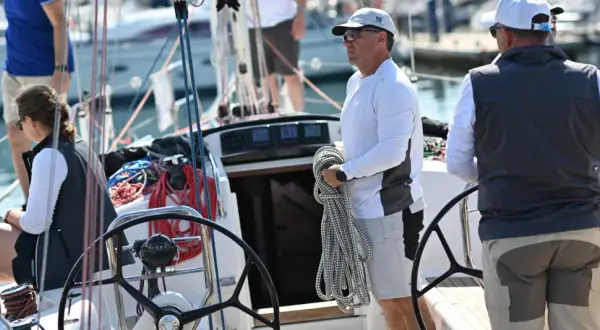 Equipaggio barca a vela pronto per la regata. Un uomo tiene una cima, una donna è seduta, mentre un altro uomo è al timone.