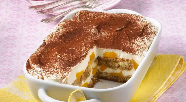 Ein cremiges Pfirsich Tiramisu, bestäubt mit Kakao, in einer weißen Auflaufform. Saftige Pfirsichstücke und Löffelbiskuits sind geschichtet.