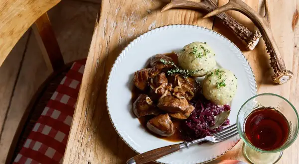 Piatto di cinghiale in dolceforte con canederli e cavolo rosso. Un bicchiere di vino rosso e un palco di cervo completano la scena rustica.