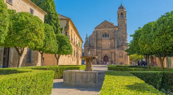 Fuente renacentista en plaza de Úbeda y Baeza, con iglesia barroca al fondo y árboles frutales.