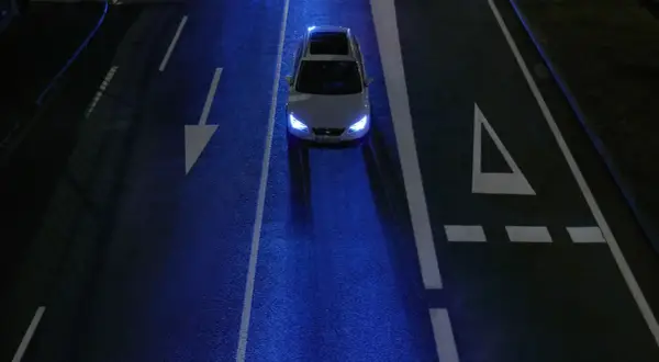 Un coche blanco con las luces encendidas, conduciendo de noche por una carretera oscura con marcas viales blancas y un reflejo azulado.