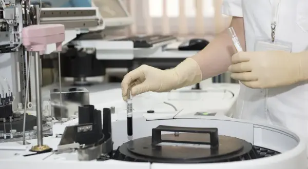 Ręka w rękawiczce umieszcza probówkę z ciemnym płynem w aparacie laboratoryjnym. To co to za badanie?