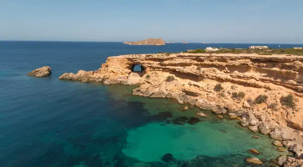 Costa rocosa con una cueva natural en Cala Comte, aguas turquesas y el mar azul profundo.