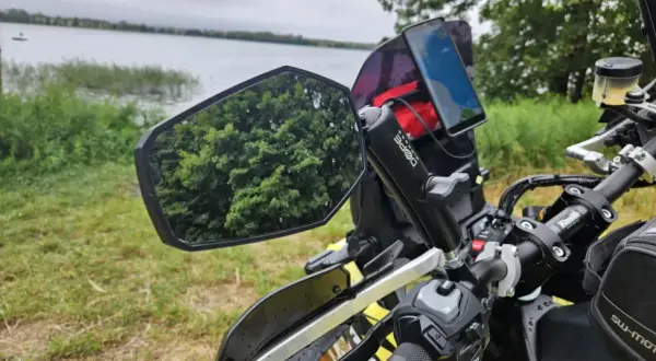 Lusterko motocyklowe z odbiciem drzew i telefonem nawigacyjnym na kierownicy, nad jeziorem.
