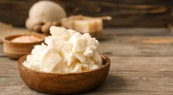 Un bol en bois rempli de beurre de karité, un ingrédient naturel pour les soins de la peau. Des noix de karité et du sel rose sont visibles en arrière-plan.