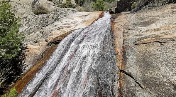 El agua desciende por la roca en la cascada del Hornillo, rodeada de vegetación y rocas graníticas bajo un cielo azul.