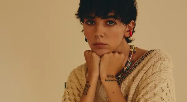 Un'ariete cantante con piercing e tatuaggi, indossa un maglione beige e una collana colorata.