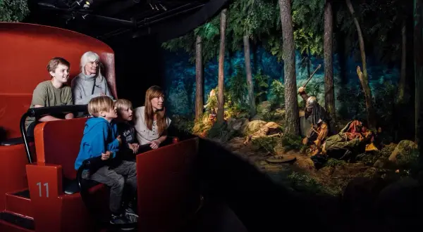 Familj i en vagn på ett vikingamuseum i Sverige, tittar på en dramatisk scen med vikingar i en skog.