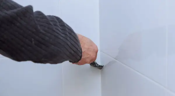 Ręka w swetrze usuwa stary silikon z narożnika białych płytek za pomocą narzędzia.