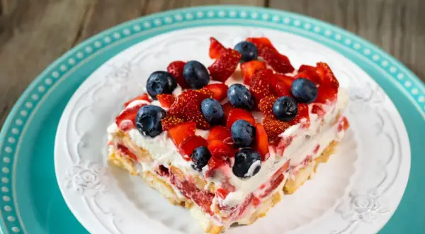 Tranche de tiramisu aux fruits rouges, fraises et myrtilles, sur une assiette blanche ornée, posée sur une assiette turquoise.