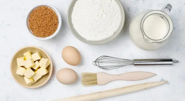 Ingredientes para hornear: harina, azúcar moreno, mantequilla en cubos, huevos, leche en jarra, batidor, brocha y rodillo de madera.