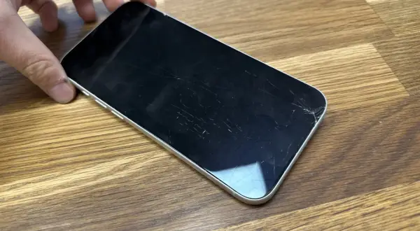 Dłoń trzyma telefon z pękniętym szkłem hartowanym. Ekran pokryty jest siecią drobnych pęknięć, szczególnie widocznych w prawym dolnym rogu.
