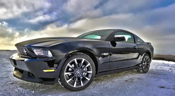 Czarny Ford Mustang GT 5.0, jeden z tych tanich sportowych aut, zaparkowany na zaśnieżonej drodze pod dramatycznym niebem.