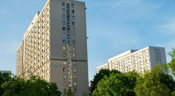 Dwa wysokie, szare bloki mieszkalne z balkonami, otoczone zielenią. W tle błękitne niebo. Taki widok to codzienność wielu lokatorów, którzy co miesiąc płacą **czynsz**.