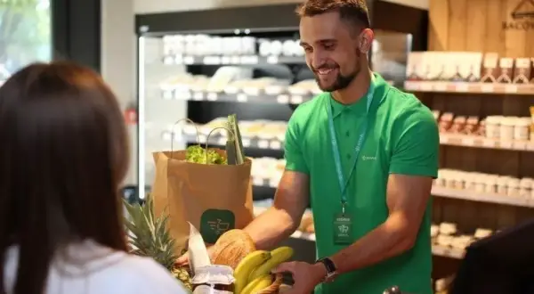 Sprzedawca w zielonej koszulce podaje klientce koszyk z zakupami w sklepie spożywczym.