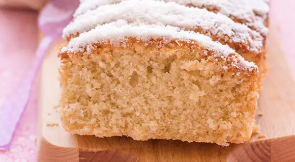 Bizcocho de coco esponjoso, cubierto con coco rallado, servido en tabla de madera.
