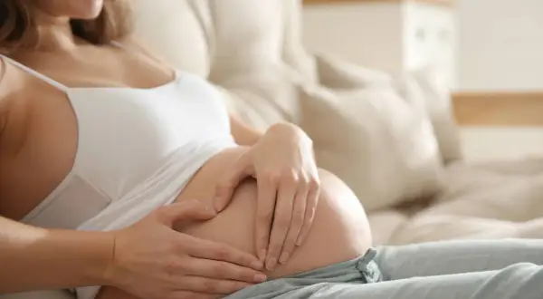 A pregnant woman rests on a couch, her hands forming a heart on her belly. She smiles, perhaps thinking about the baby, boy or girl, and the early pregnancy insomnia.