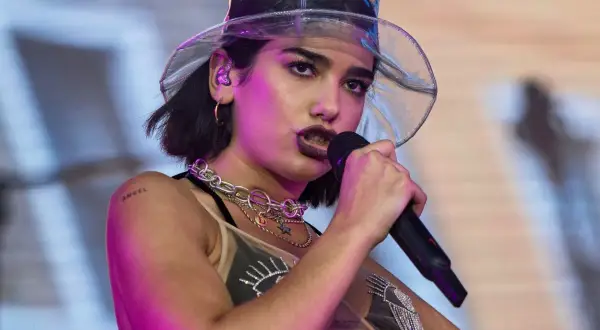 Dua Lipa w przezroczystym kapeluszu śpiewa do mikrofonu na koncercie.