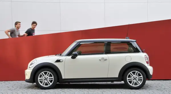 Biały Mini Cooper r56 z czarnym dachem stoi na bruku. Dwóch mężczyzn biegnie za nim na tle czerwono-białej ściany.
