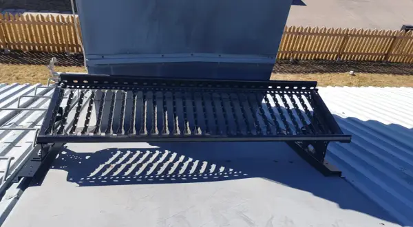 En takbrygga framför en stor ventilationsanläggning, liknande en skorsten. Solen kastar ett mönstrat skuggspel.