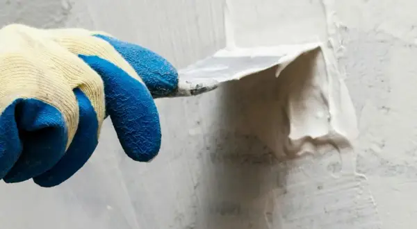 Hand med blå gummihandske spacklar en vägg utomhus. Snickerispackel utomhus används för att jämna till ytan.