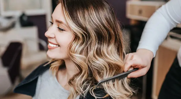 Mujer sonríe mientras estilista peina su cabello con tinte sobre mechas rubias, creando un look radiante.