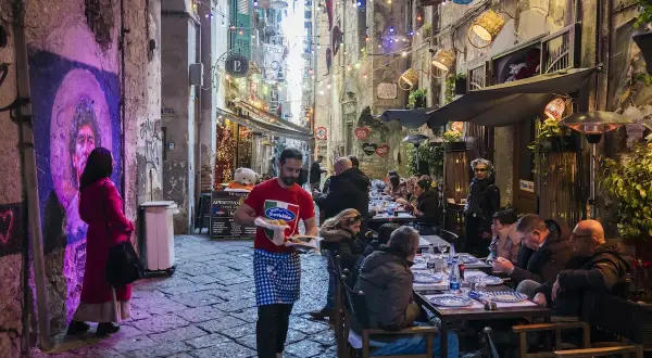 Un vicolo napoletano illuminato da luci colorate, con gente seduta ai tavoli di un locale. Un cameriere porta del cibo.
