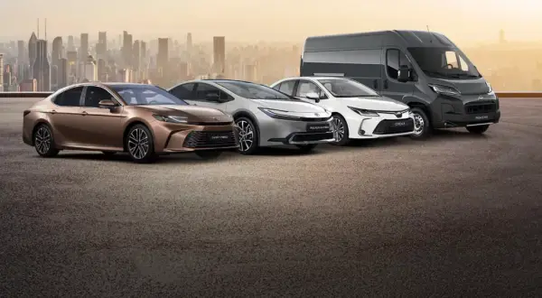 Różnorodne modele Toyota: brązowa Camry Hybrid, srebrny Prius Plug-in Hybrid, biała Corolla i szary Proace Max na tle miasta.