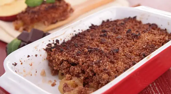 Crumble cacao appena sfornato in una teglia rossa, con mele e cioccolato a lato.