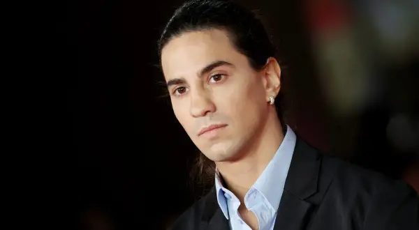 Ritratto di Tedua, rapper italiano, con capelli lunghi legati e un orecchino scintillante. La sua età è difficile da definire, ma emana un'aura di maturità artistica.
