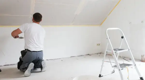 Mann kniet und spachtelt Deckenfugen, um Rigipsplatten zu kleben. Eine Leiter und ein Eimer stehen bereit.