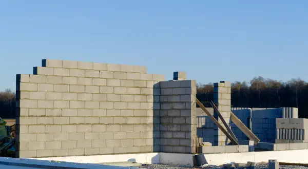Budowa domu w toku. Wznoszona jest ściana nośna z bloczków betonowych, widoczne fundamenty i materiały budowlane.