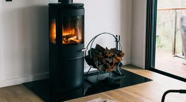 Moderner Holzofen mit brennendem Feuer, daneben ein Korb mit Holzscheiten. Ein gemütlicher Anblick, der an ein Verbot von Holzöfen denken lässt.