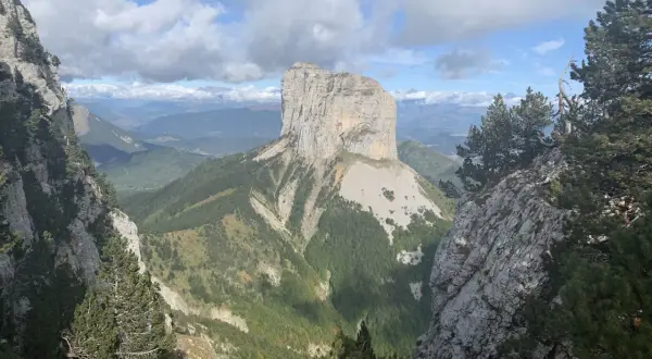 Majestätischer Mont Aiguille ragt aus grünen Tälern empor, umgeben von Felsen und Kiefern unter einem dramatischen Wolkenhimmel.