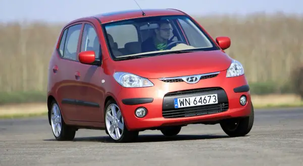 Czerwony Hyundai i10 na drodze. Kierowca w okularach i zielonym szaliku. Wiele pozytywnych hyundai i10 opinie potwierdza jego miejski charakter.