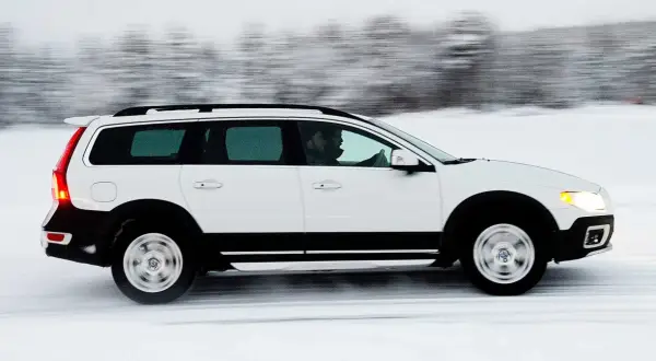 Vit Volvo XC70 kör på en snöig väg, med en förare bakom ratten. Bilen är i rörelse, vilket skapar en suddig bakgrund av träd.