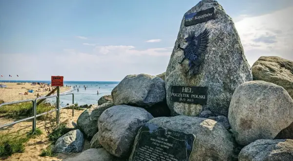 Pomnik "Początek Polski" na plaży na cyplu helskim. W tle widać morze i plażowiczów.