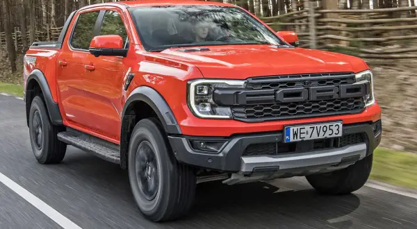 Pomarańczowy Ford Raptor jedzie po drodze w lesie. Sprawdź aktualną ford raptor cena i możliwości tego potężnego pickupa.