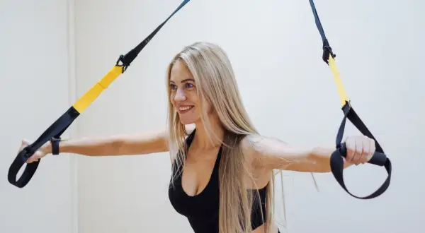 Uśmiechnięta blondynka ćwiczy z taśmami TRX, realizując swój plan treningowy na siłownię.