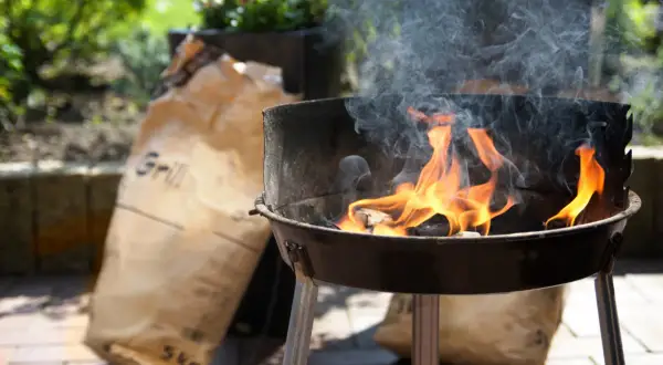 Płonący grill z workami węgla drzewnego w tle.