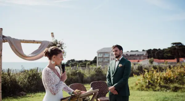La mariée, en robe blanche, parle au micro lors de la trame cérémonie laïque. Le marié, en costume vert, l'écoute attentivement.