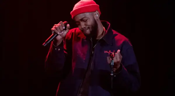 Ant Clemons, wearing a red beanie and dark jacket, sings passionately into a microphone on a dimly lit stage.