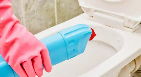 Mano con guanto rosa usa detergente per pulire il water incrostato.